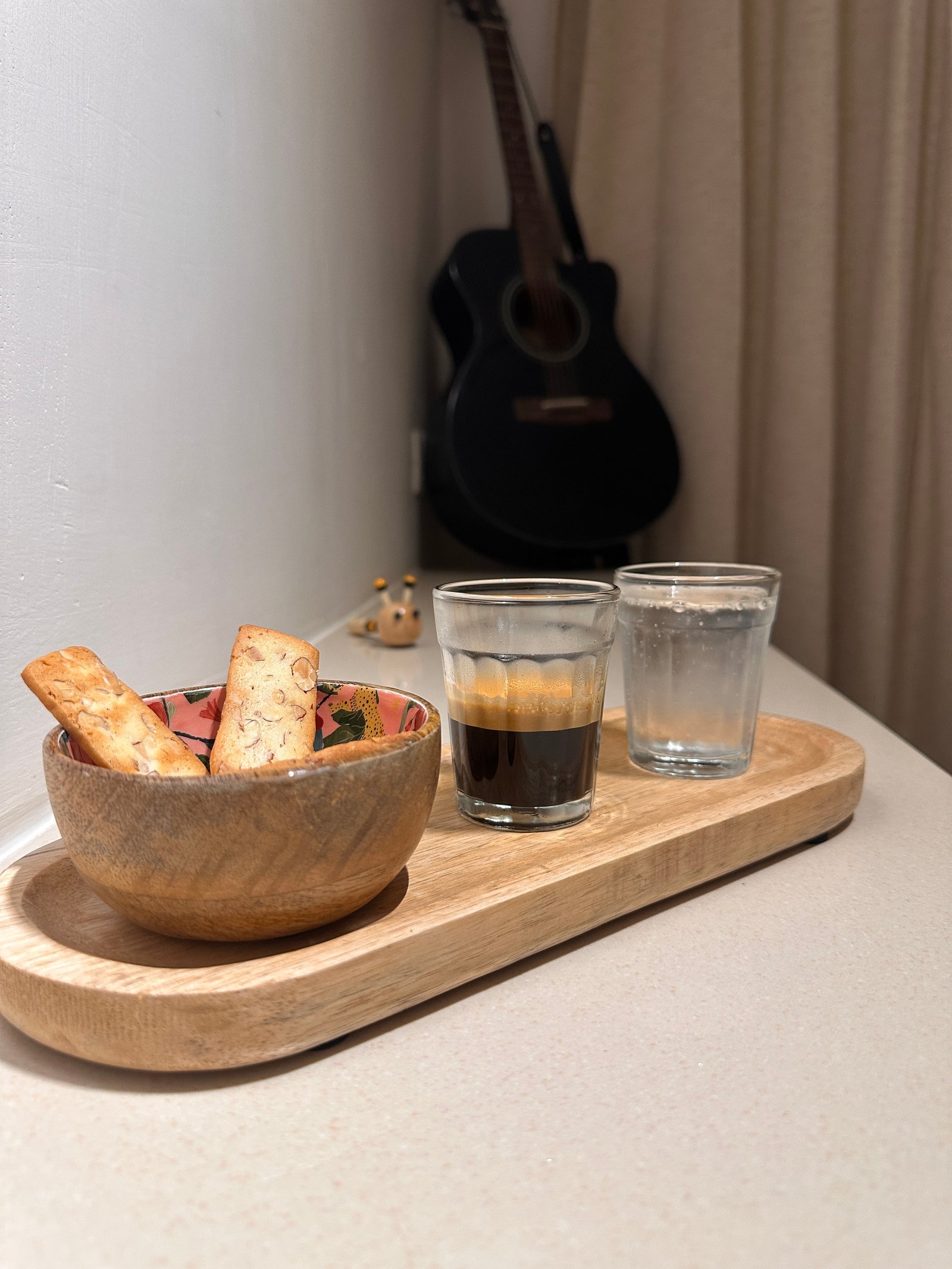 A wooden tray with a bowl of almond biscuits, a glass of espresso and a glass of sparkling water, with a guitar in the background.